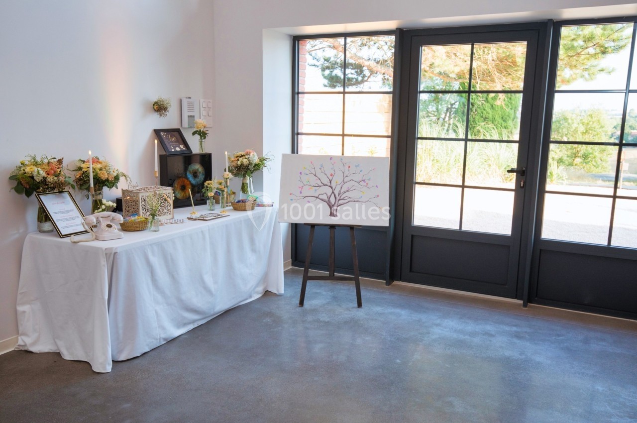 Table décorée avec des fleurs, cadres et bougies, à côté d'un tableau représentant un arbre, dans une pièce lumineuse.