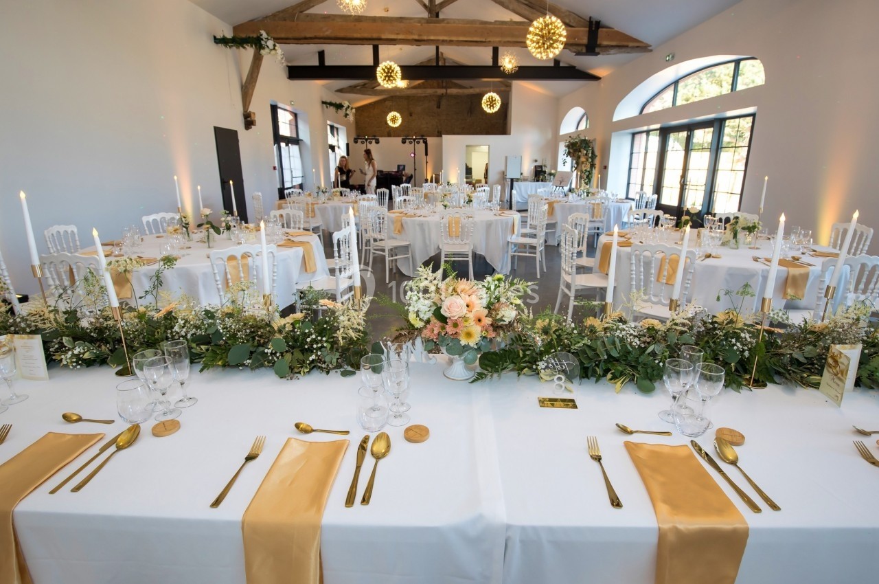 Salle de réception décorée pour un mariage avec tables dressées, nappes blanches, chemins de table dorés et compositions…