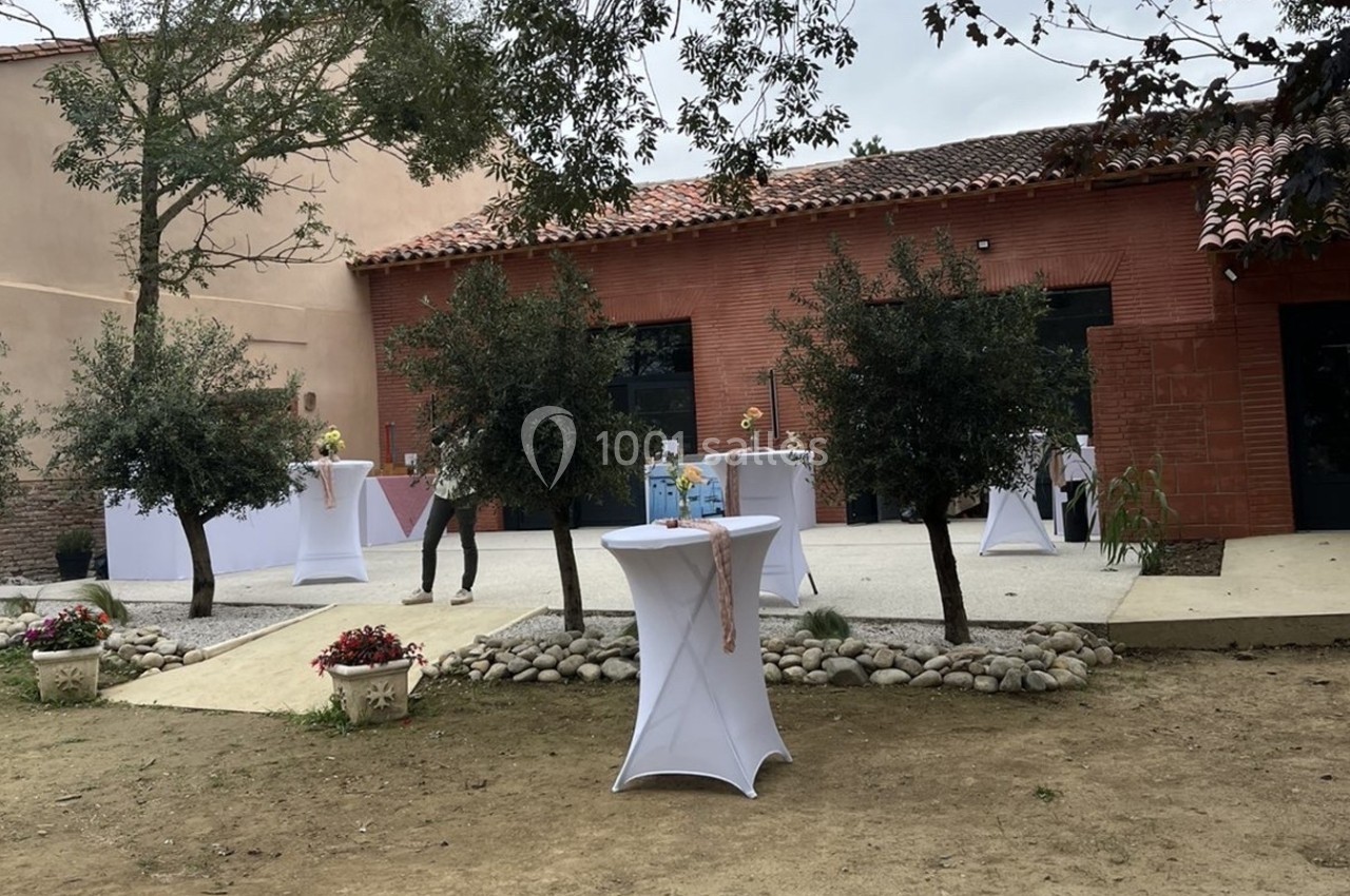 Terrasse extérieure avec tables hautes décorées, entourée d'arbres et de bâtiments en briques rouges.