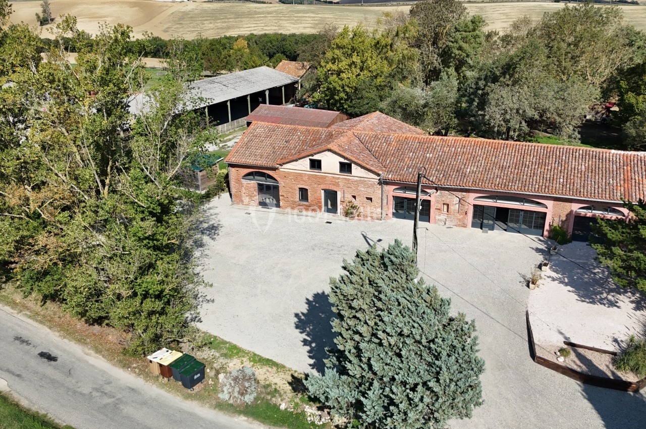 Vue aérienne d'un bâtiment en briques avec toit en tuiles, entouré d'arbres et situé près d'une route de campagne.
