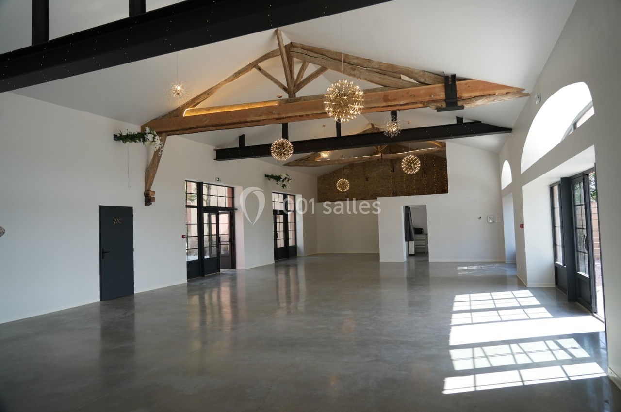 Salle lumineuse avec poutres apparentes, sol en béton poli, grandes fenêtres et suspensions décoratives.