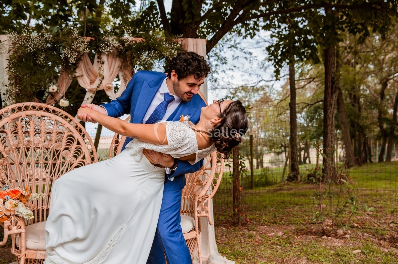 Un couple en tenue de mariage danse devant une arche décorée de fleurs dans un cadre extérieur boisé.