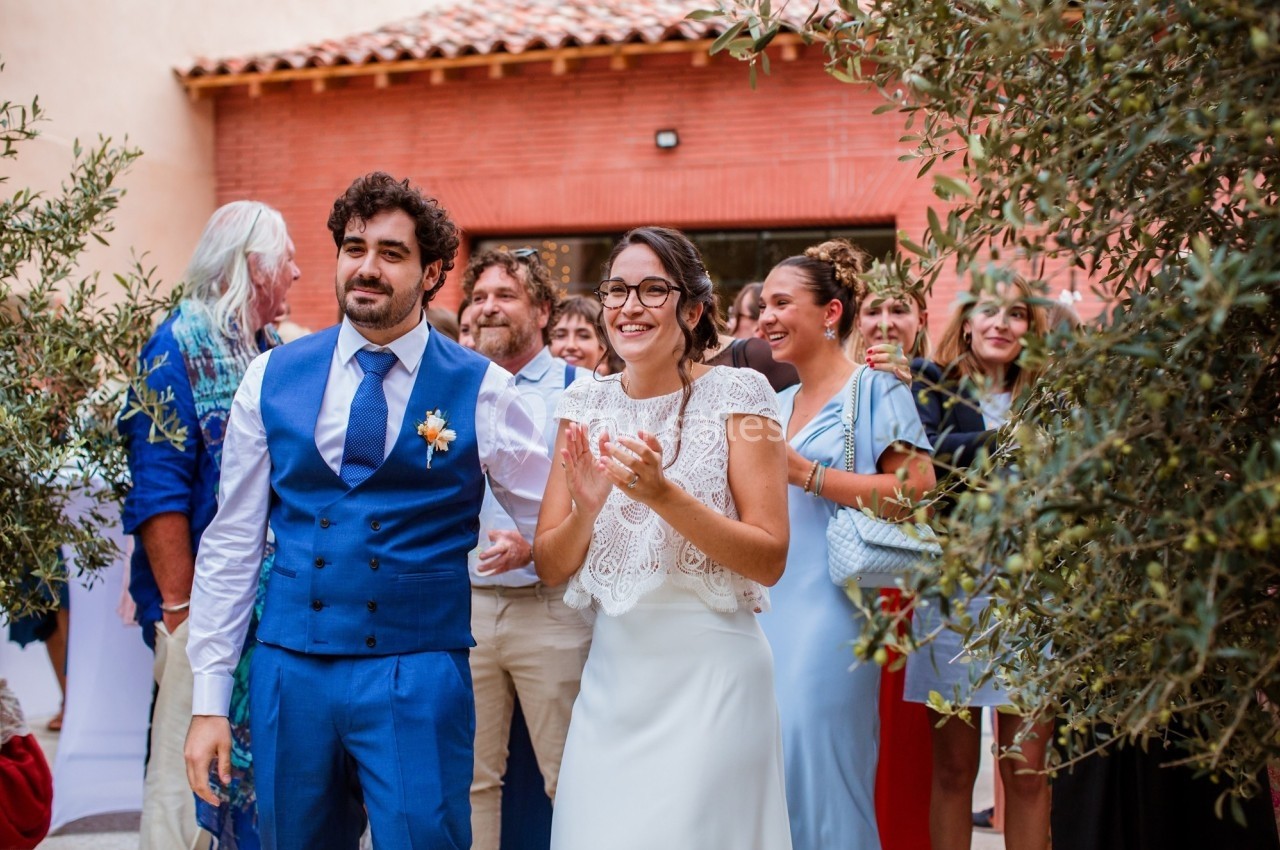 Un couple souriant, entouré d'invités, se tient debout dans un cadre extérieur avec des oliviers.