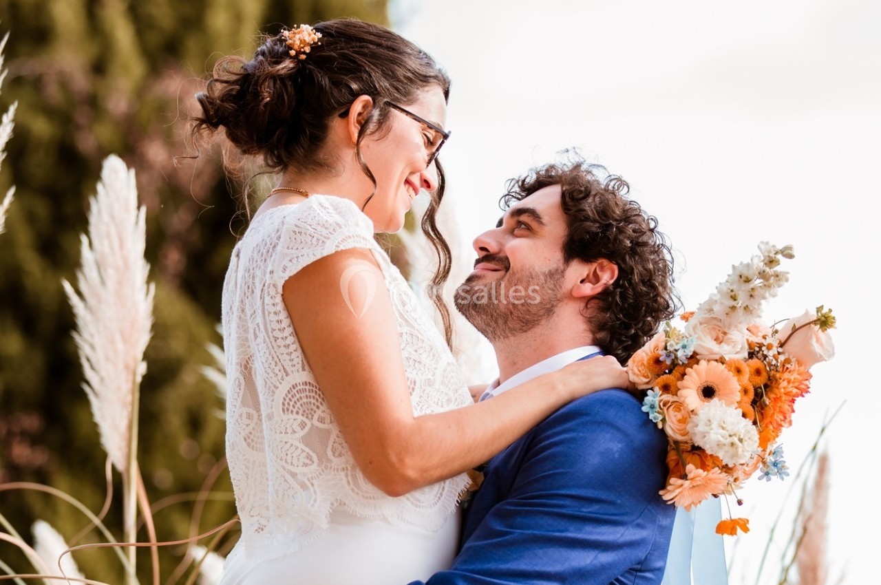 Une mariée en robe blanche et un marié en costume bleu se regardent avec tendresse, bouquet coloré en arrière-plan.