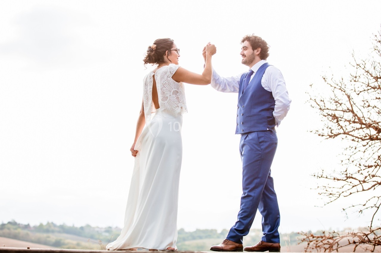 Un couple en tenue de mariage danse en extérieur sous un ciel lumineux, avec un paysage naturel en arrière-plan.