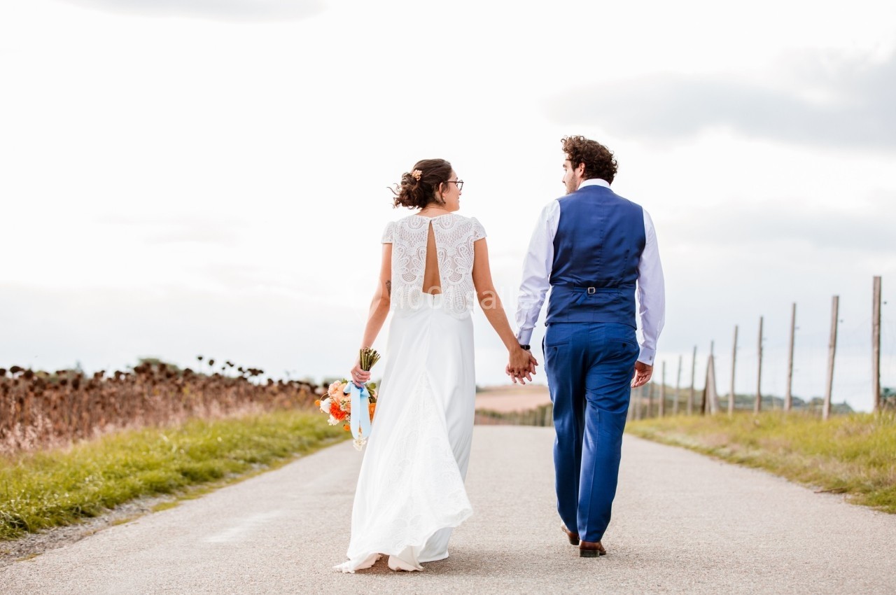 Un couple en tenue de mariage marche main dans la main sur une route de campagne par temps clair.