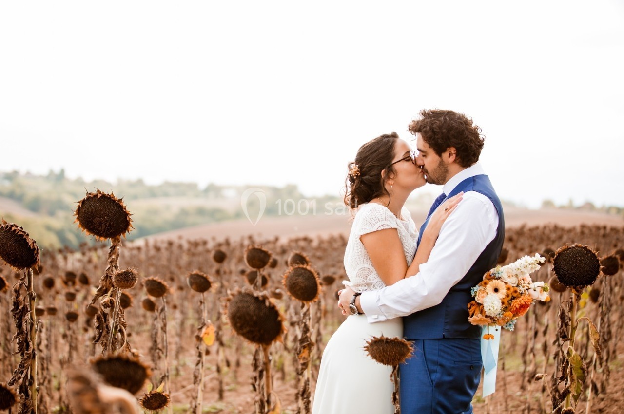 Un couple s'embrasse dans un champ de tournesols fanés, la mariée tenant un bouquet de fleurs.