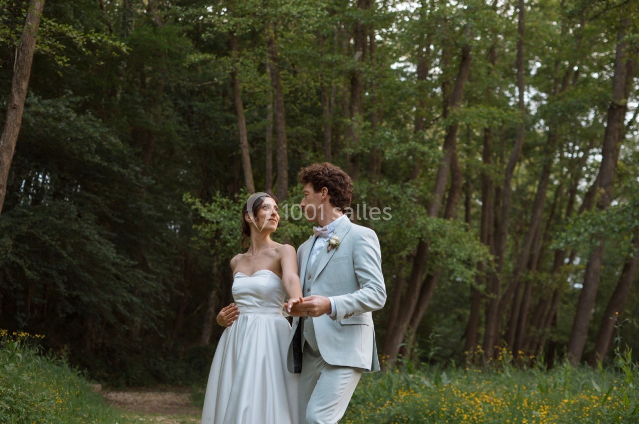 Un couple en tenue de mariage marche dans une forêt verdoyante, échangeant un regard complice.