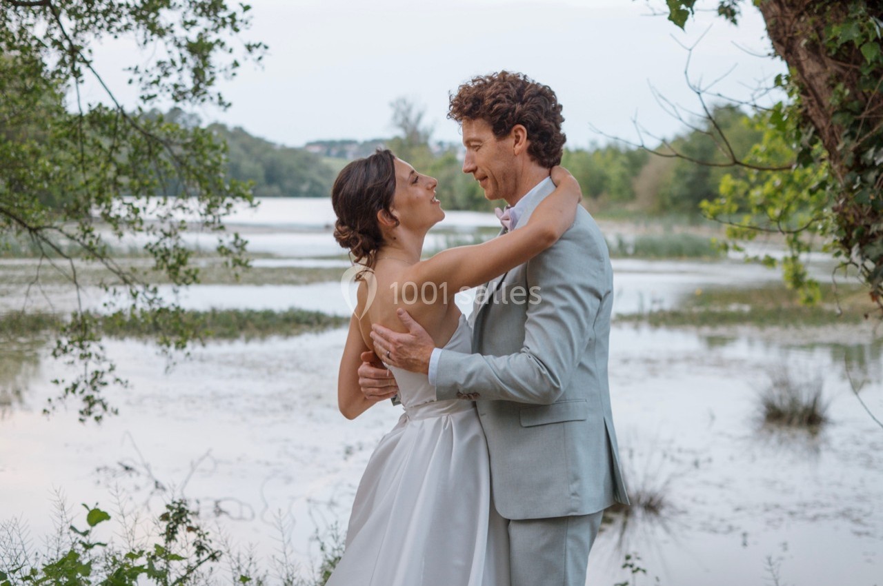 Un couple en tenue de mariage s'enlace près d'un lac entouré de végétation.