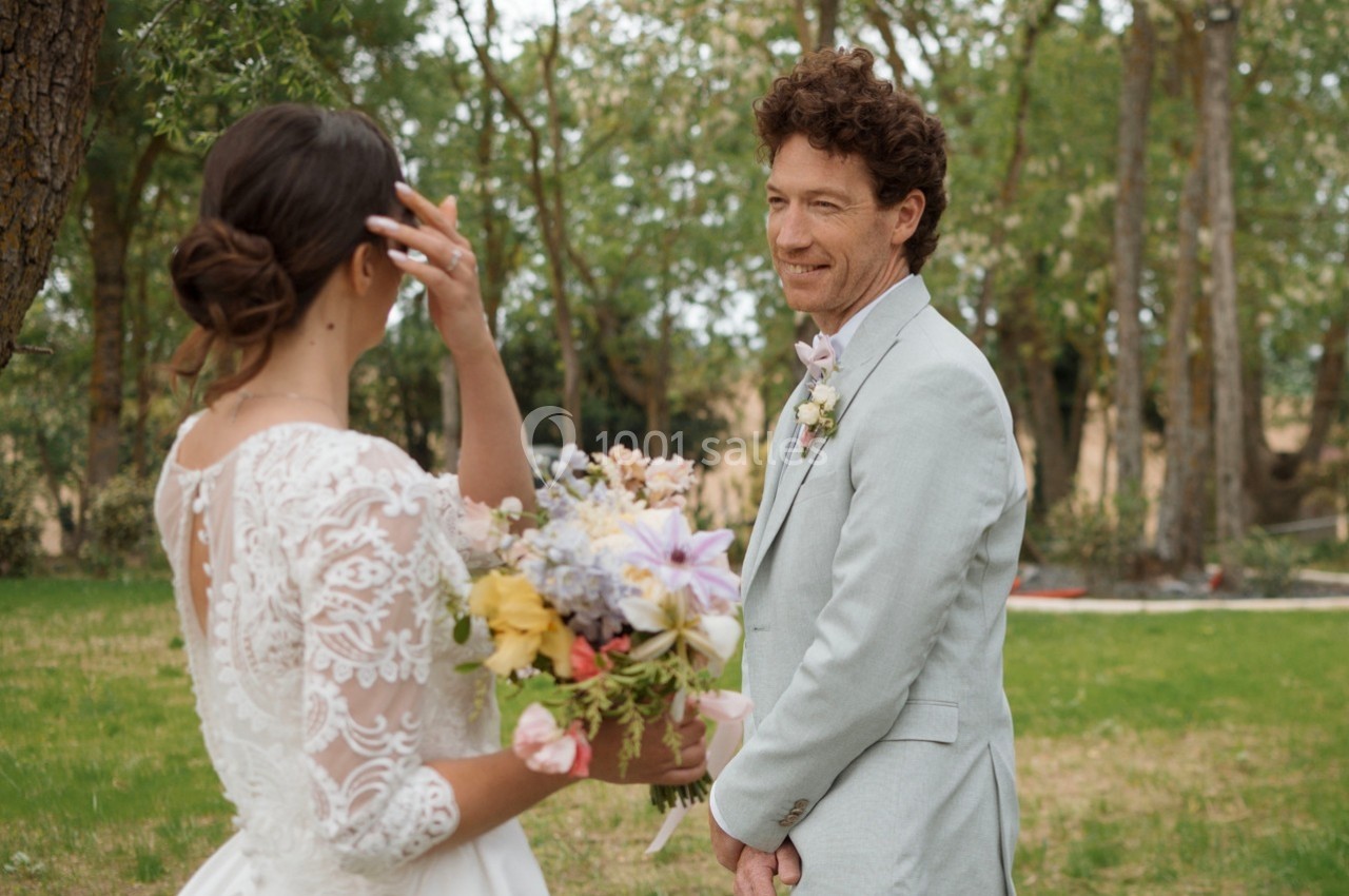 Une mariée en robe blanche tient un bouquet coloré face à un homme en costume clair dans un jardin verdoyant.