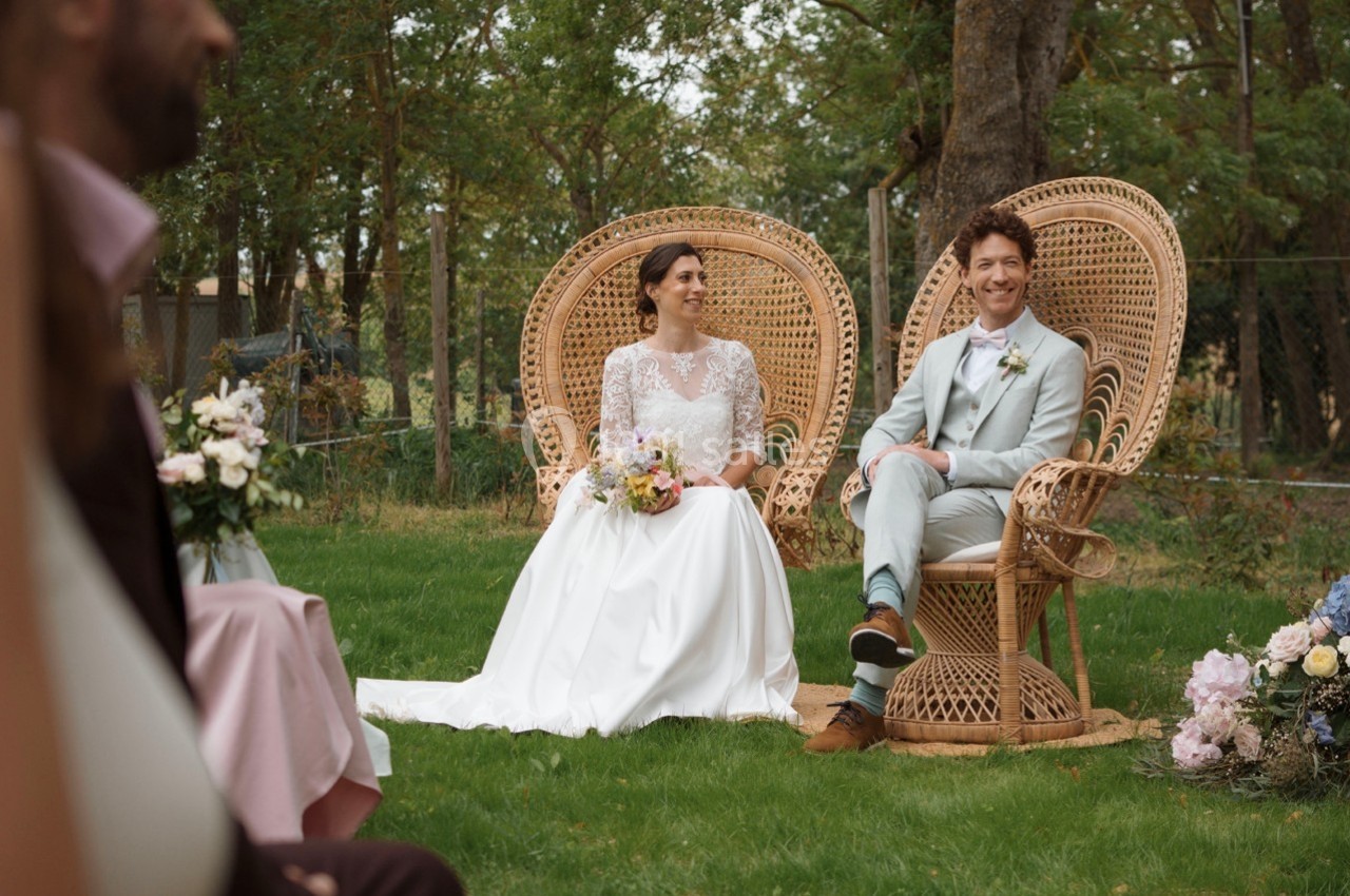 Un couple assis sur des fauteuils en osier lors d'une cérémonie de mariage en extérieur, entouré de verdure.