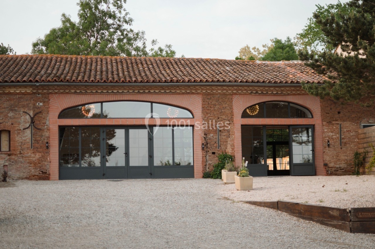 Façade d'un bâtiment en briques avec grandes baies vitrées, entouré de végétation et d'une cour en gravier.