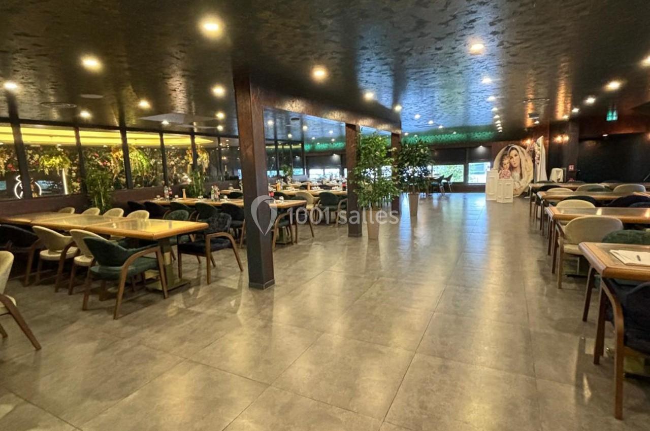 Salle de restaurant spacieuse avec tables en bois, chaises rembourrées, plantes décoratives et éclairage chaleureux.
