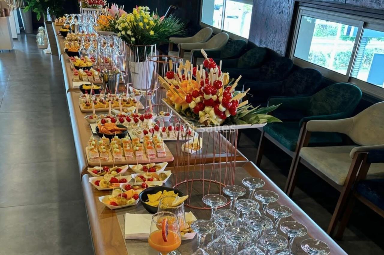 Buffet avec verrines, brochettes de fruits, toasts et verres disposés sur une longue table dans une salle lumineuse.