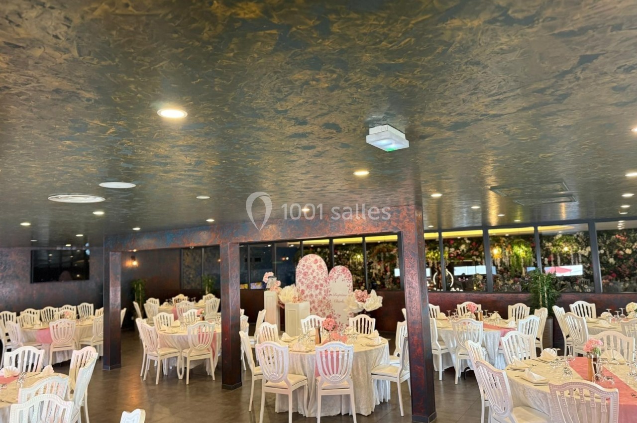 Salle de réception décorée avec des tables rondes, nappes roses et chaises blanches, éclairée par des lumières au plafond.