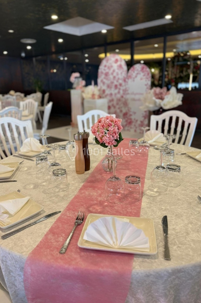 Table décorée pour un événement avec nappe blanche, chemin de table rose, vaisselle élégante et bouquet de fleurs roses.