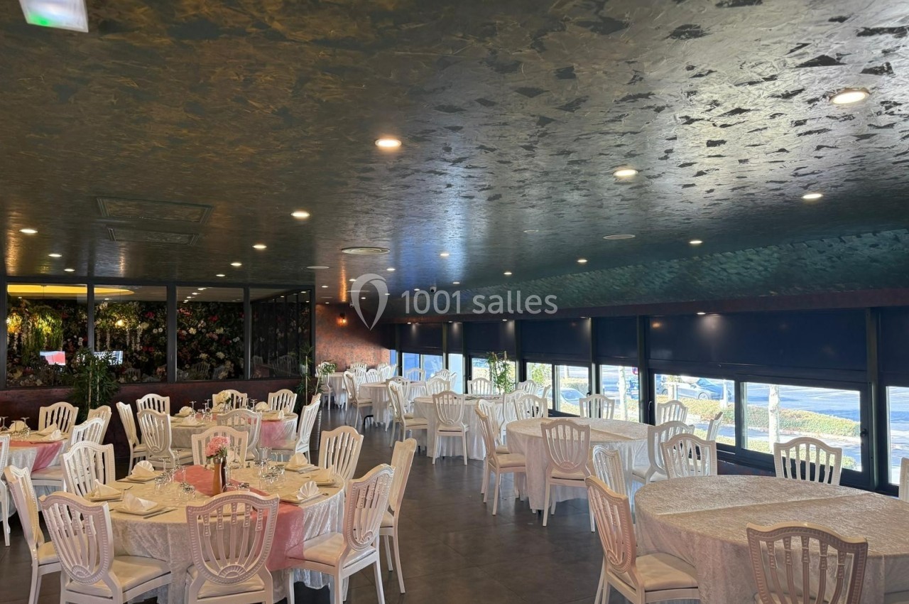 Salle de réception lumineuse avec des tables rondes dressées, chaises blanches et grandes baies vitrées.