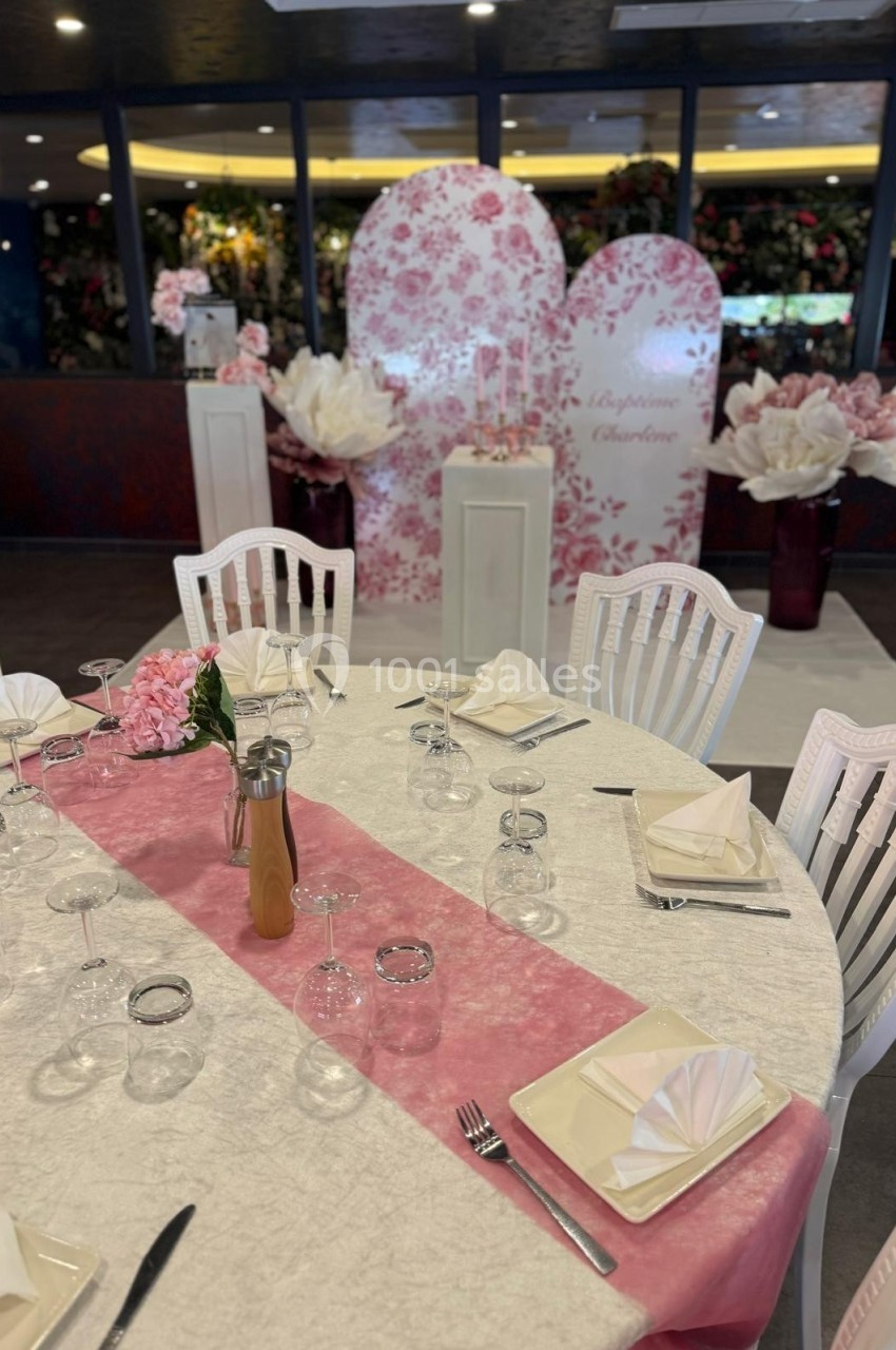 Table ronde élégamment dressée avec nappe blanche, chemin de table rose, serviettes pliées et décor floral en arrière-plan.