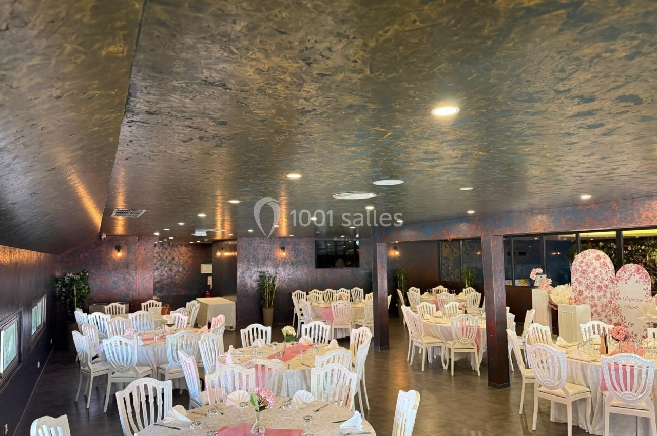 Salle de réception décorée avec des tables rondes dressées, chaises blanches et ambiance lumineuse chaleureuse.
