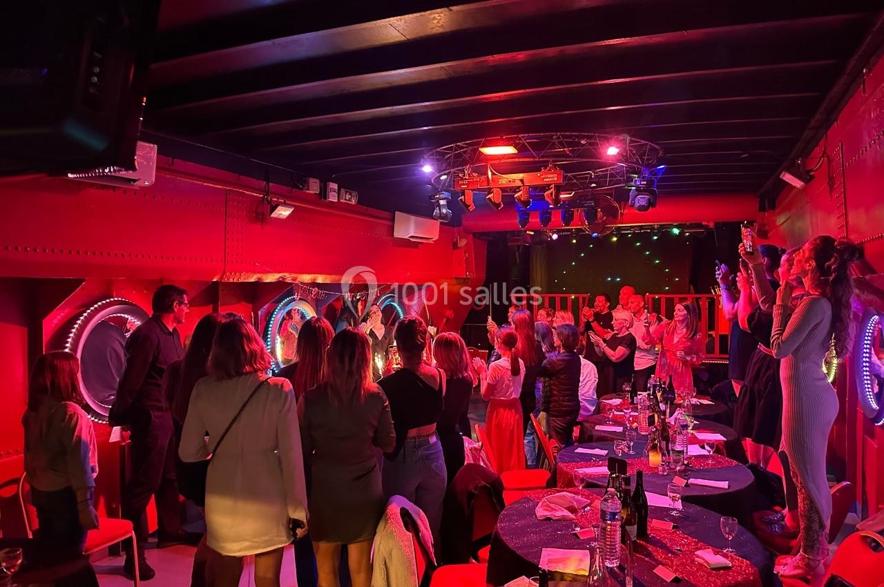 Salle animée avec des tables dressées, des convives debout applaudissant sous des lumières rouges et violettes.