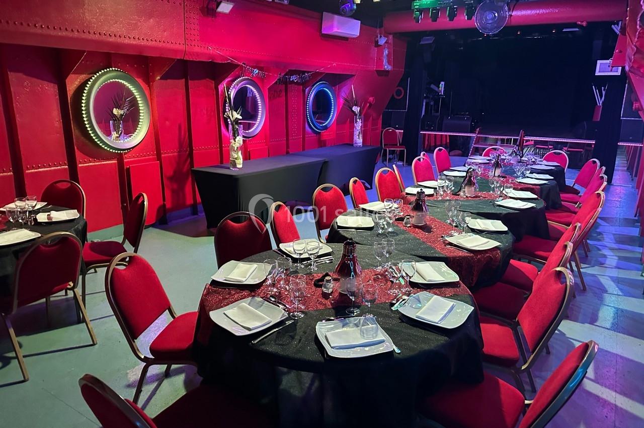 Salle décorée avec des tables rondes dressées, nappes noires et chaises rouges, éclairage tamisé et ambiance festive.