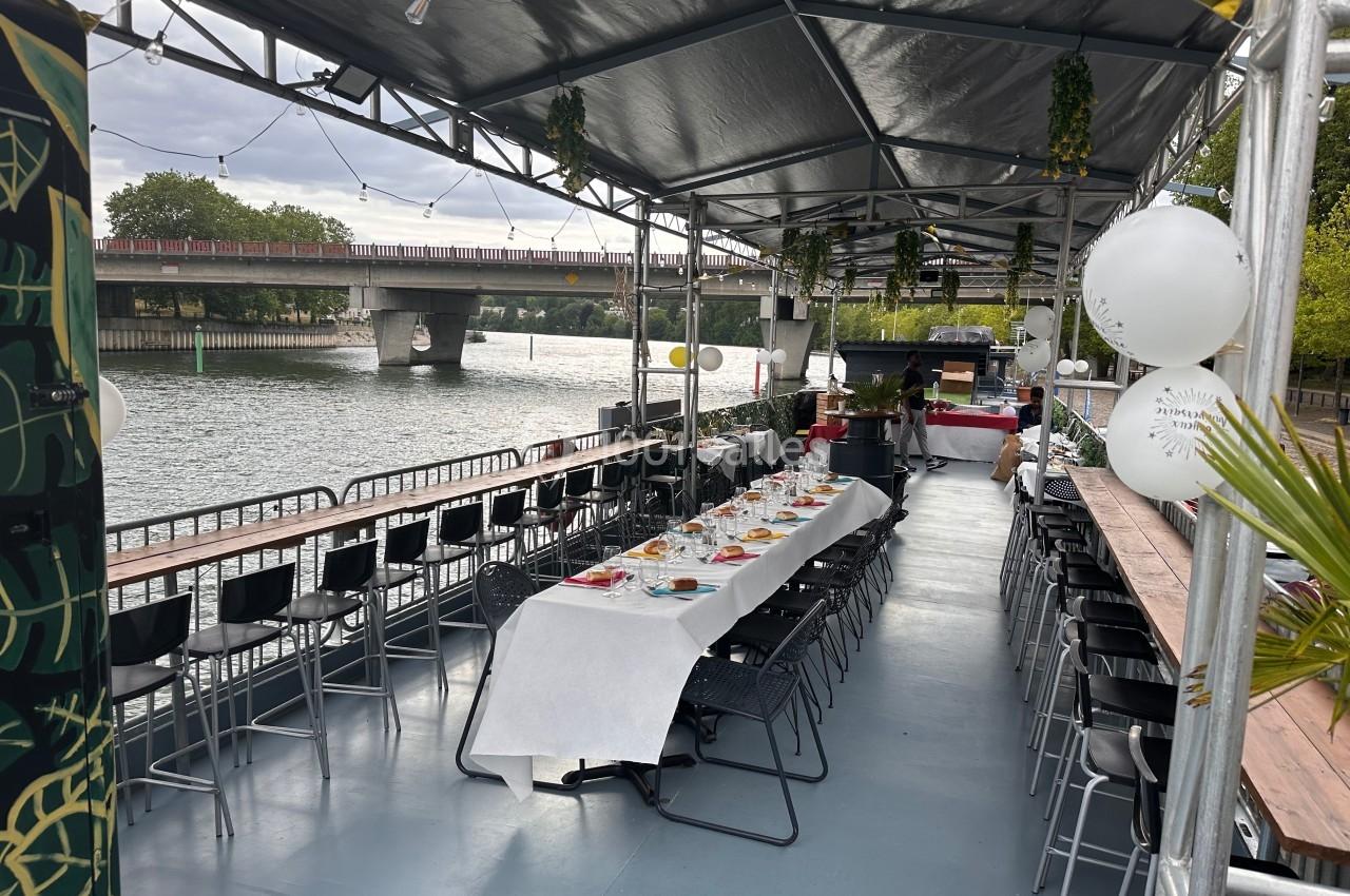 Tables dressées sous une tente sur une péniche, avec vue sur un fleuve et un pont en arrière-plan.
