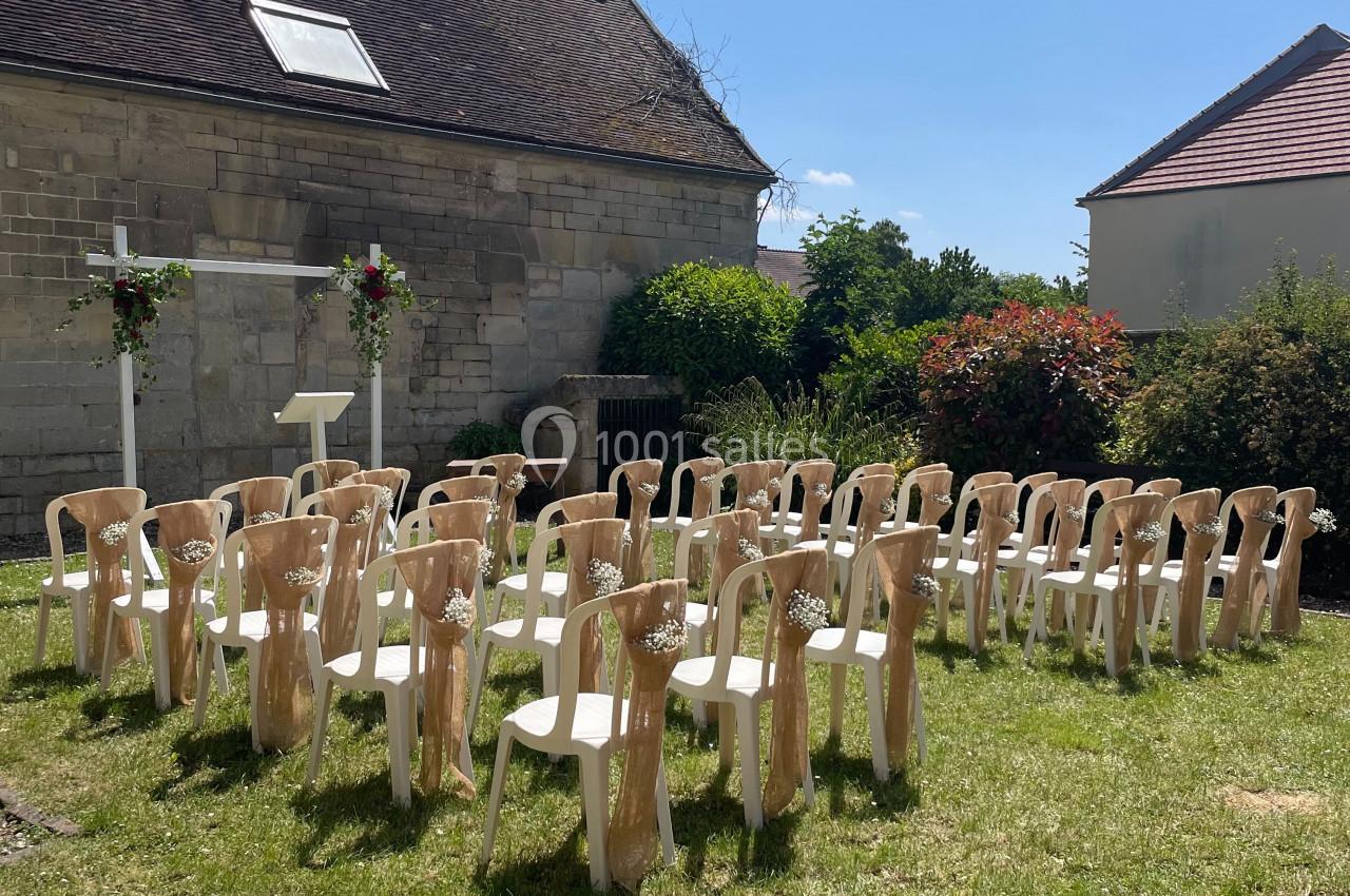Chaises blanches décorées disposées en rangées sur une pelouse, face à une arche fleurie dans un jardin ensoleillé.