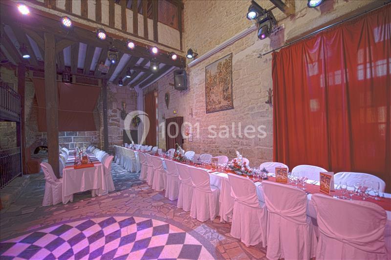 Salle de réception avec de longues tables dressées, chaises blanches, murs en pierre et rideaux rouges.