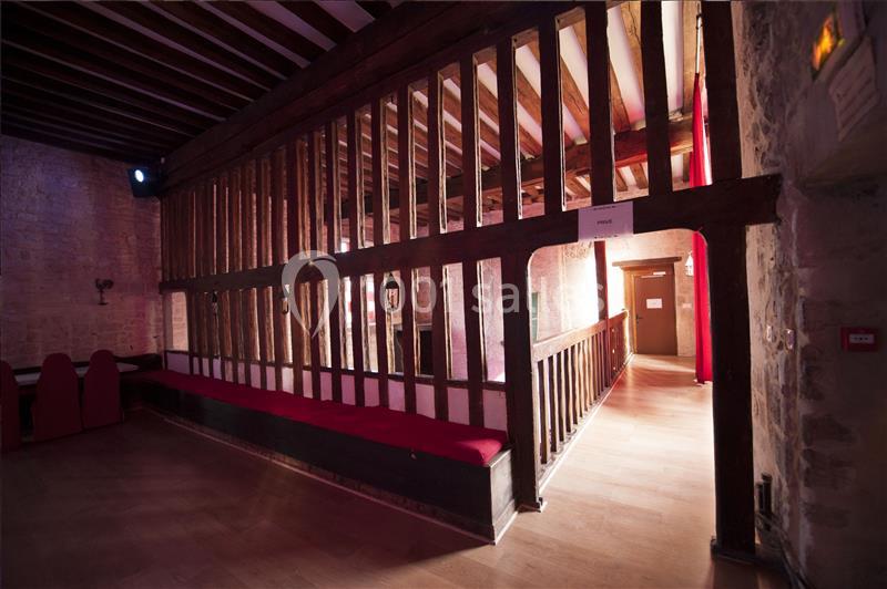 Vue intérieure d'une salle avec des murs en pierre, une structure en bois, un banc rouge et un couloir éclairé.