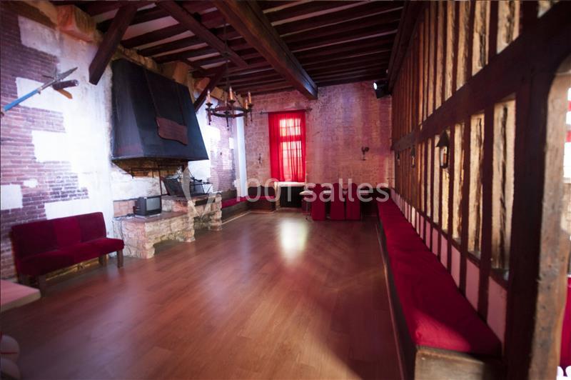 Salle rustique avec poutres apparentes, cheminée en pierre, murs en briques et sièges rouges alignés.