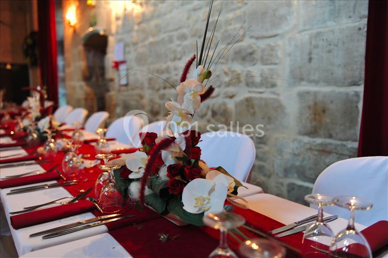 Table décorée avec des nappes rouges, des fleurs blanches et rouges, et des verres alignés dans une salle en pierre.