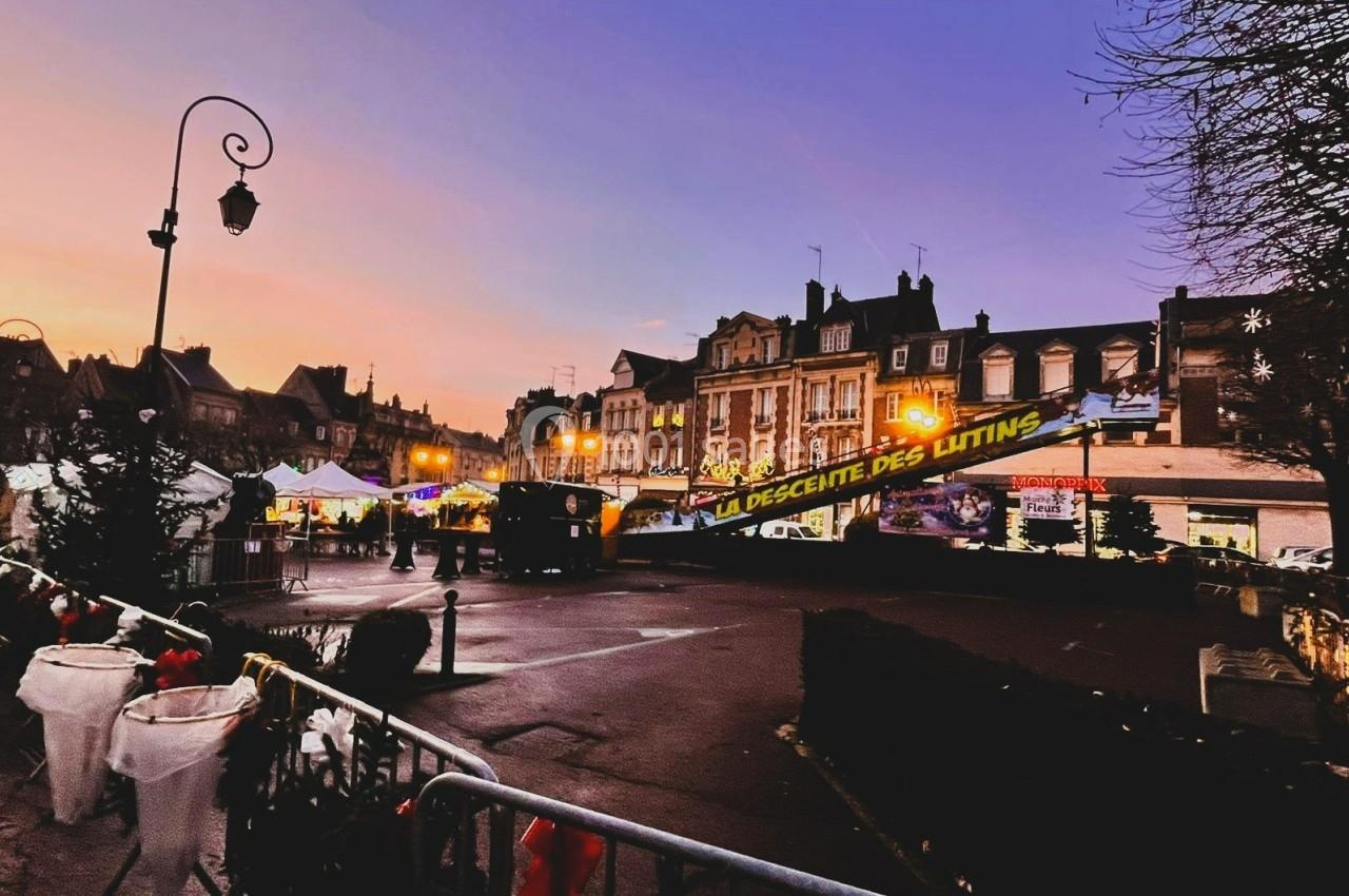 Place animée au crépuscule avec des stands illuminés, des décorations festives et une bannière ’La Descente des Lutins’.