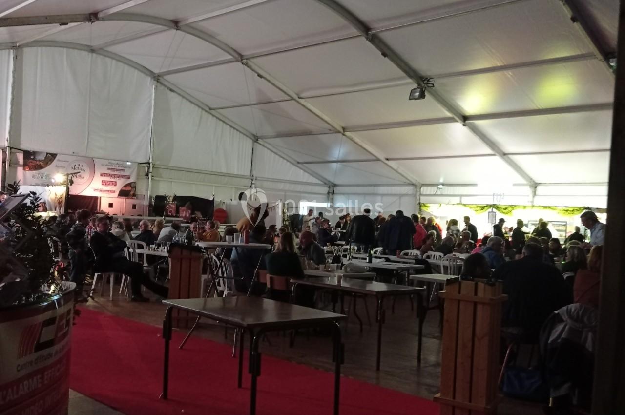 Intérieur d'un grand chapiteau avec des tables, des chaises et des personnes assises ou debout dans une ambiance animée.