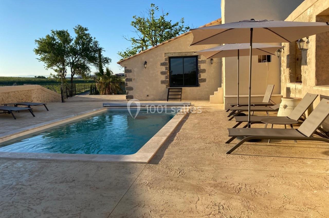 Piscine extérieure entourée de chaises longues et parasols, située près d'une maison en pierre sous un ciel dégagé.