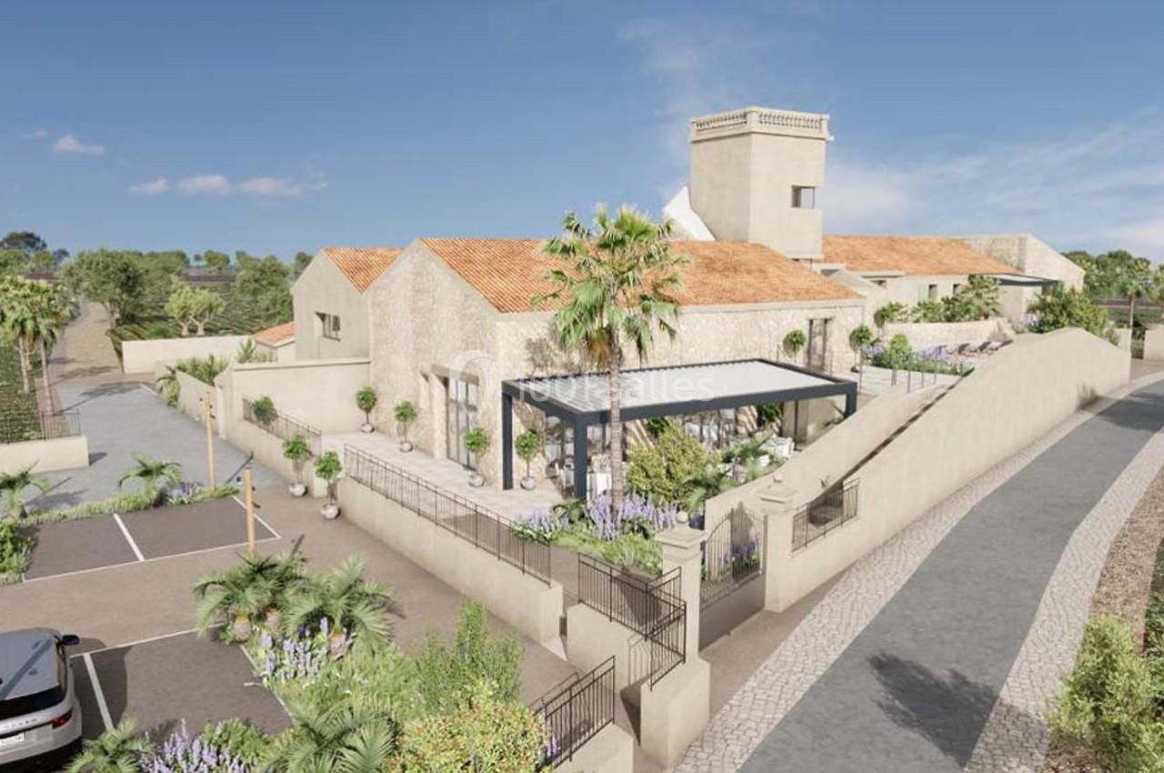 Vue d'un bâtiment en pierre avec toit en tuiles, entouré de végétation, d'une terrasse et d'un parking adjacent.
