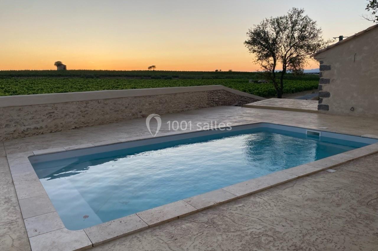 Piscine extérieure entourée de dalles en pierre, avec vue sur un champ et un coucher de soleil à l'horizon.