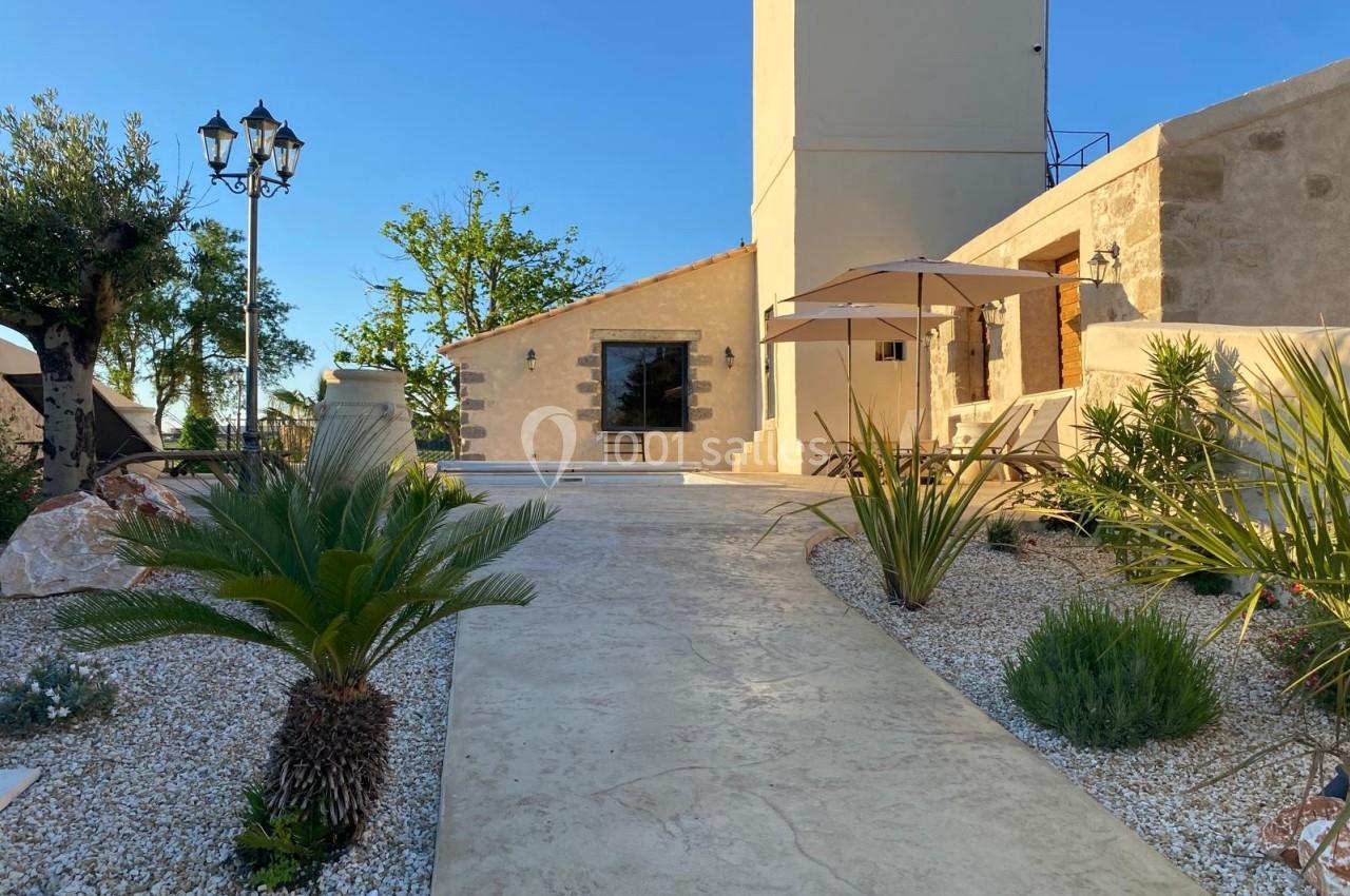 Allée en pierre bordée de plantes menant à une maison en pierre avec terrasse et parasol, sous un ciel dégagé.