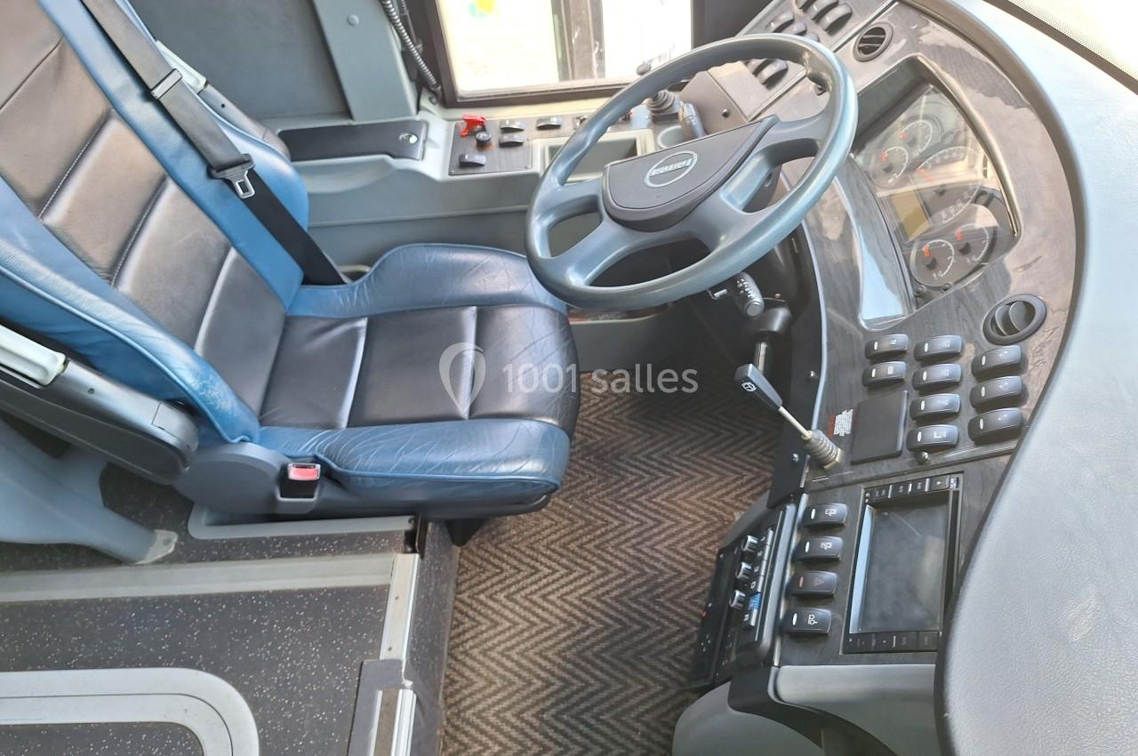 Poste de conduite d'un bus avec siège en cuir bleu, volant, tableau de bord et commandes visibles.