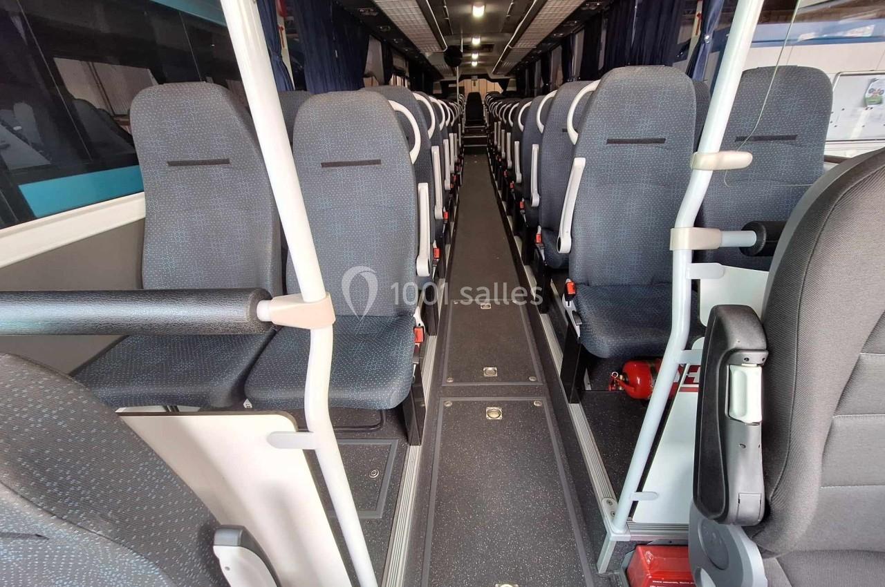 Intérieur d'un autocar avec rangées de sièges vides, allée centrale et barres de maintien visibles.