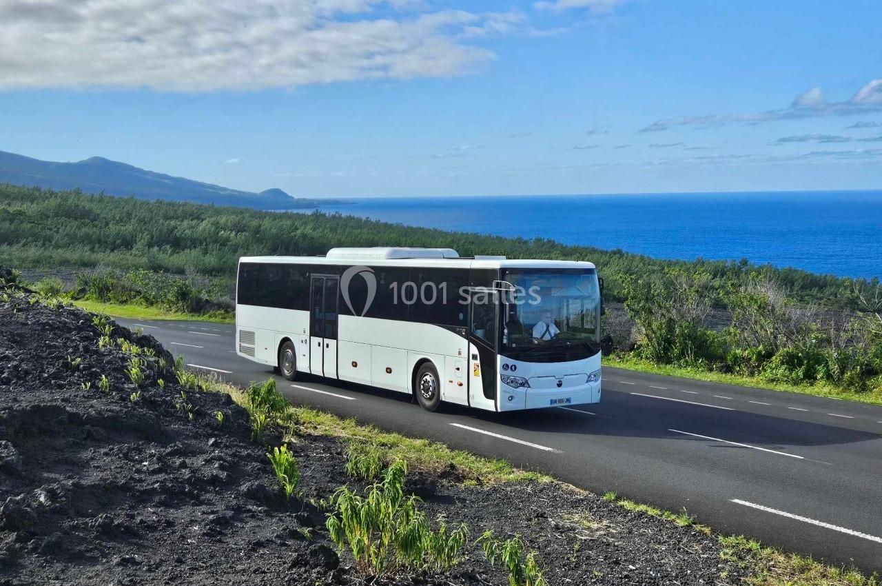 Autocar blanc circulant sur une route bordée de végétation, avec vue sur l'océan et des collines en arrière-plan.