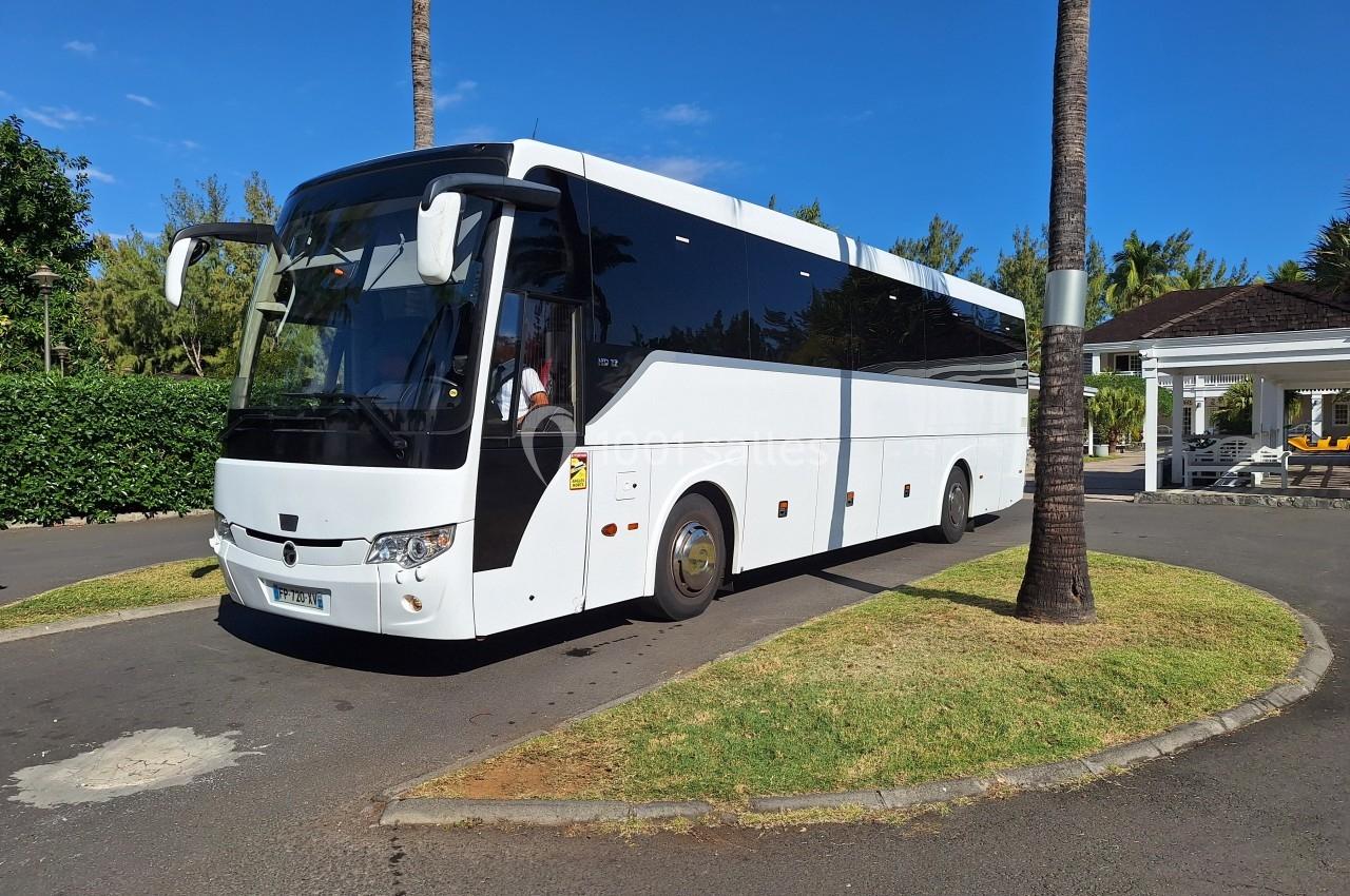 Autocar blanc stationné sur une allée pavée entourée de palmiers et de végétation sous un ciel bleu.