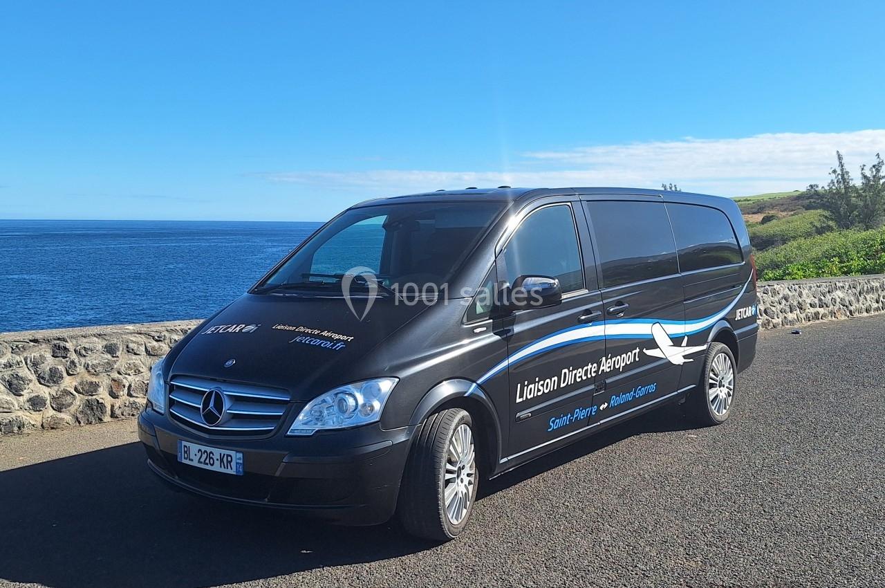 Minibus noir stationné près d'un muret en bord de mer, avec un paysage côtier en arrière-plan.