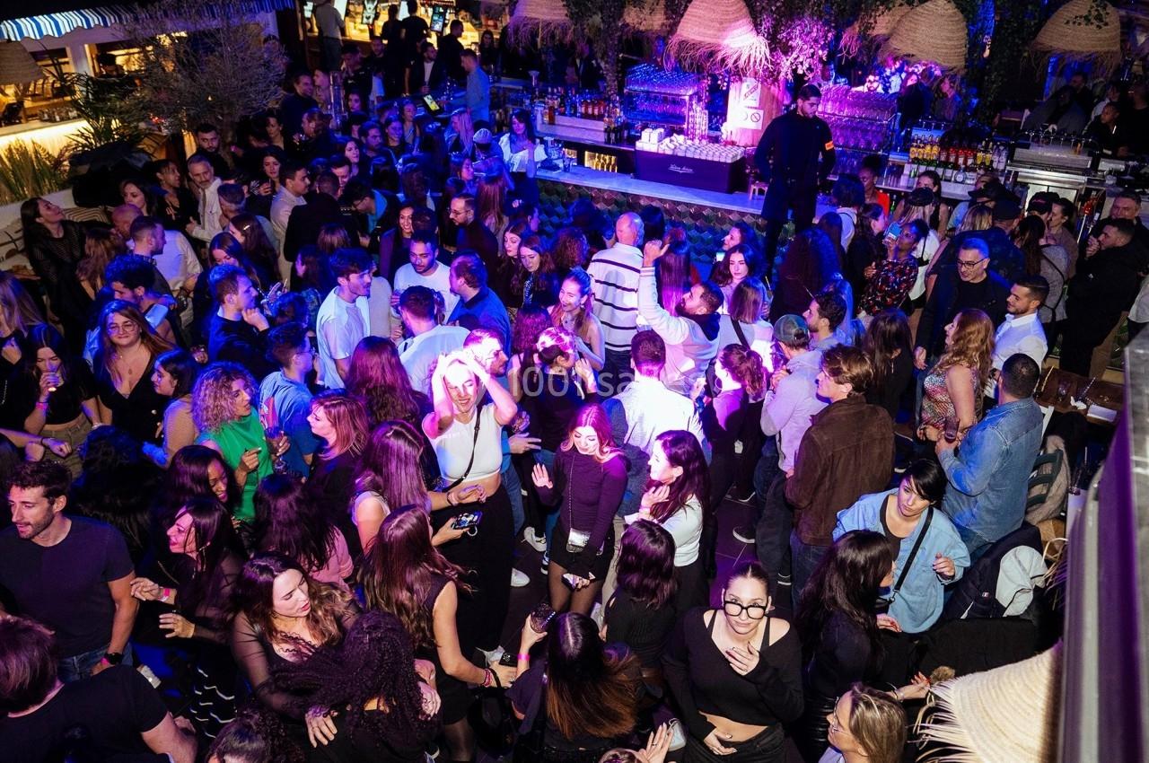 Foule de personnes dans un bar ou club animé, éclairé par des lumières colorées, avec un comptoir en arrière-plan.