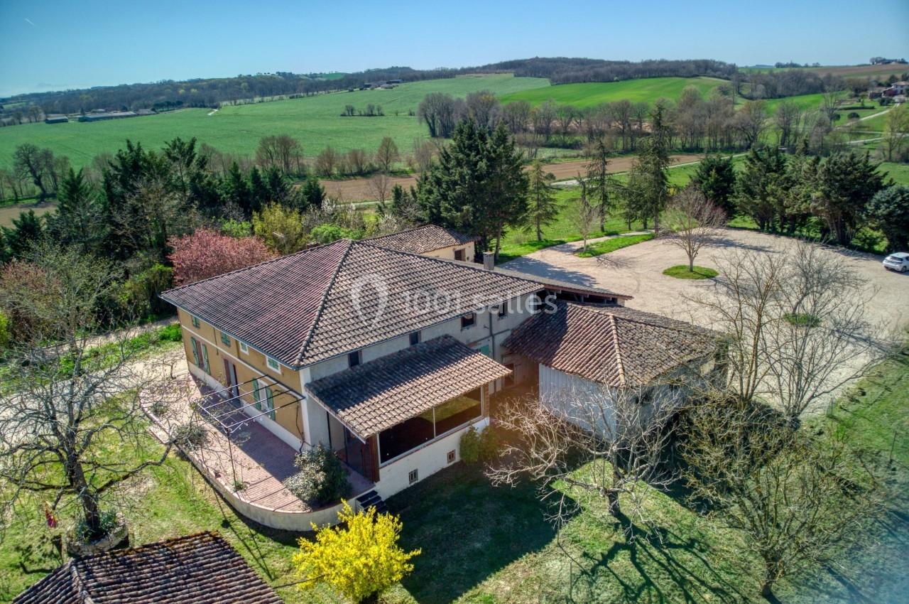 Vue aérienne d'une maison avec toit en tuiles, entourée de verdure et de champs sous un ciel dégagé.
