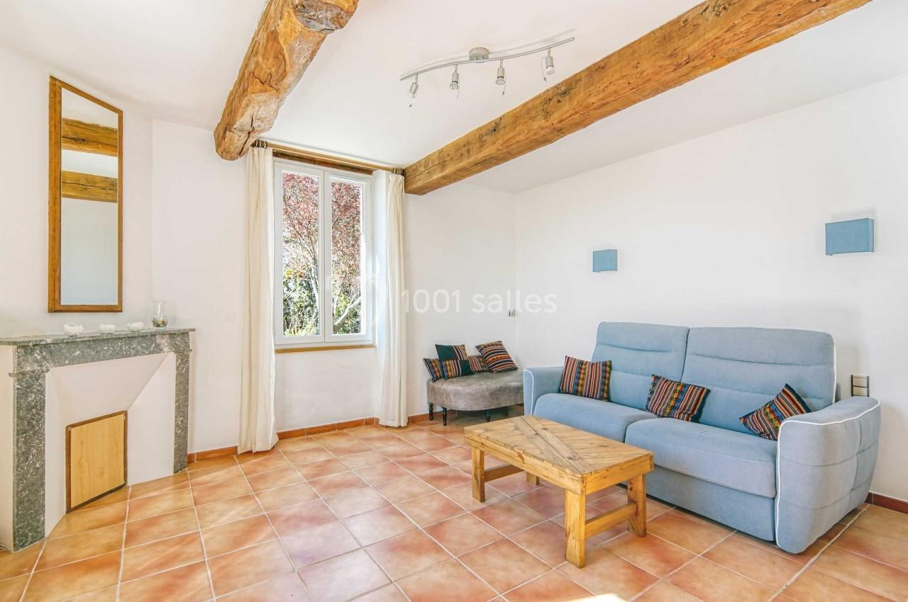 Salon lumineux avec poutres apparentes, canapé bleu, table basse en bois, cheminée et fenêtre donnant sur un arbre.