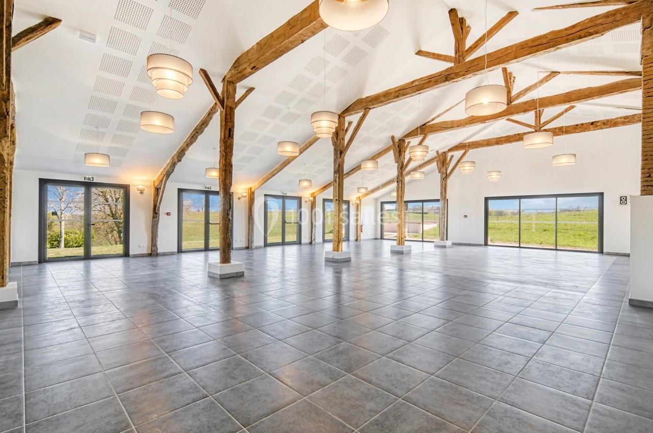 Salle spacieuse avec poutres apparentes, carrelage gris et grandes baies vitrées donnant sur un paysage verdoyant.