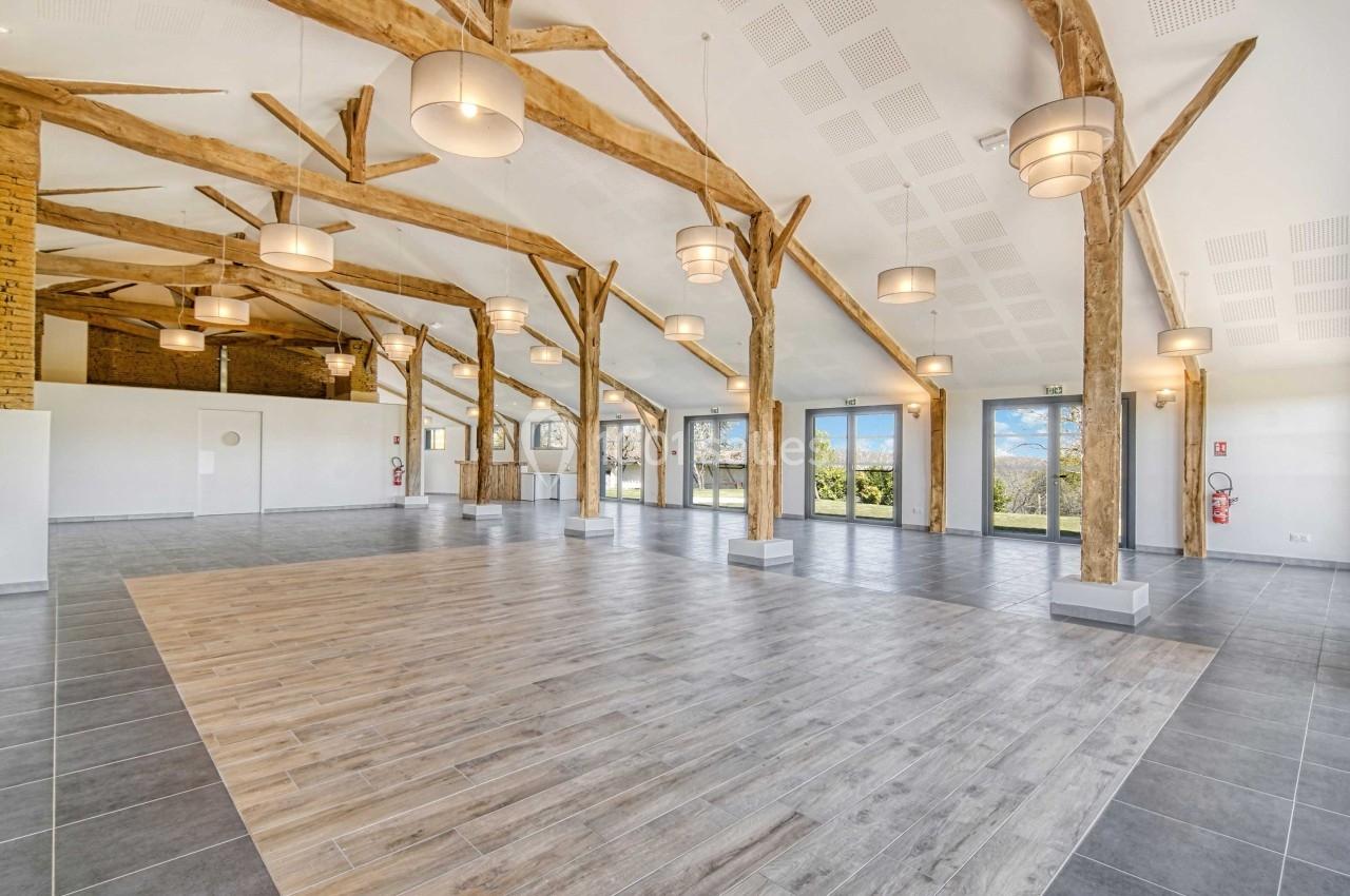 Salle spacieuse avec poutres en bois apparentes, sol en parquet et carrelage, grandes baies vitrées donnant sur un jardin.