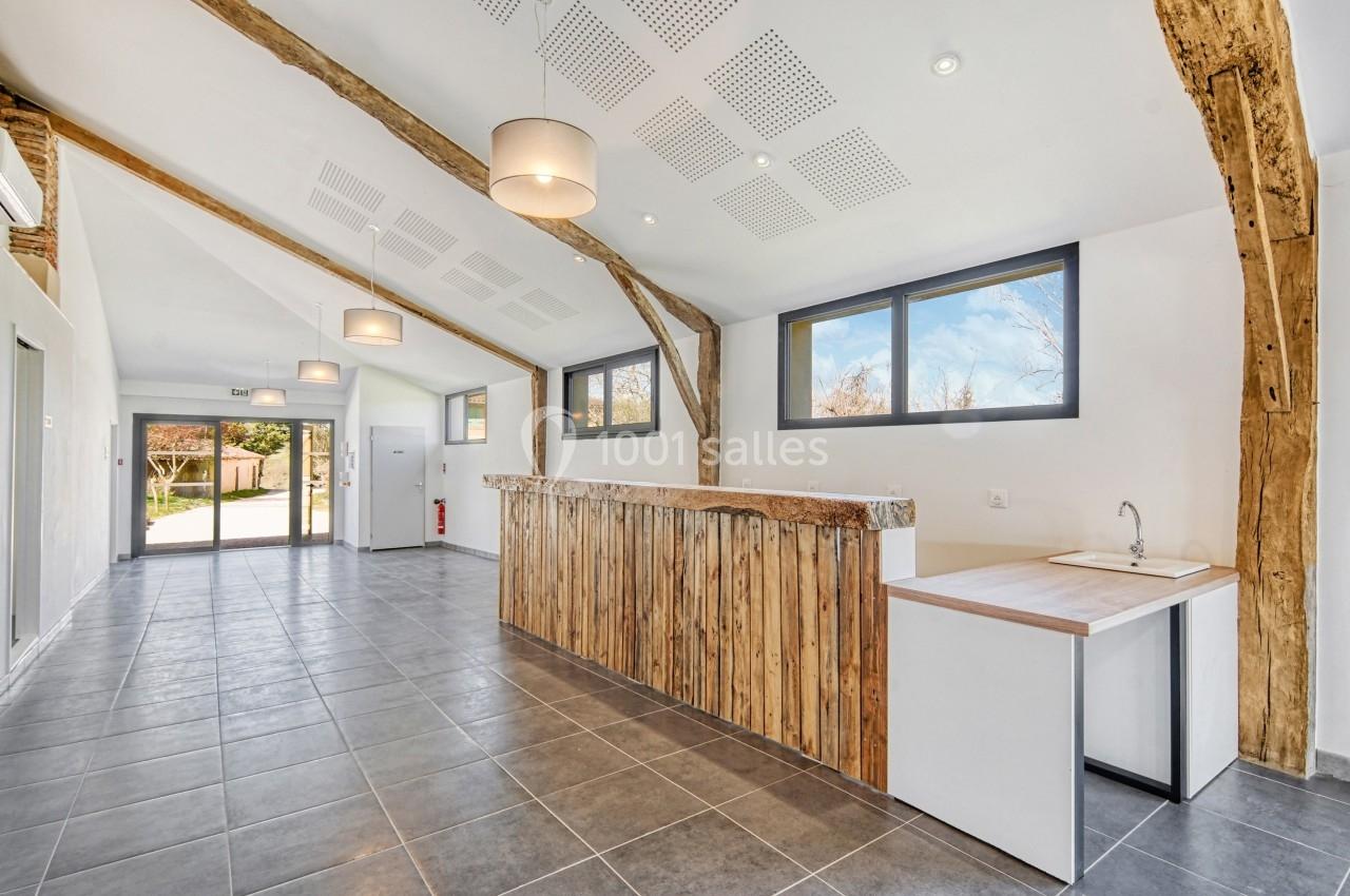 Salle lumineuse avec comptoir en bois, poutres apparentes, sol en carrelage gris et grandes fenêtres donnant sur l'extérieur.