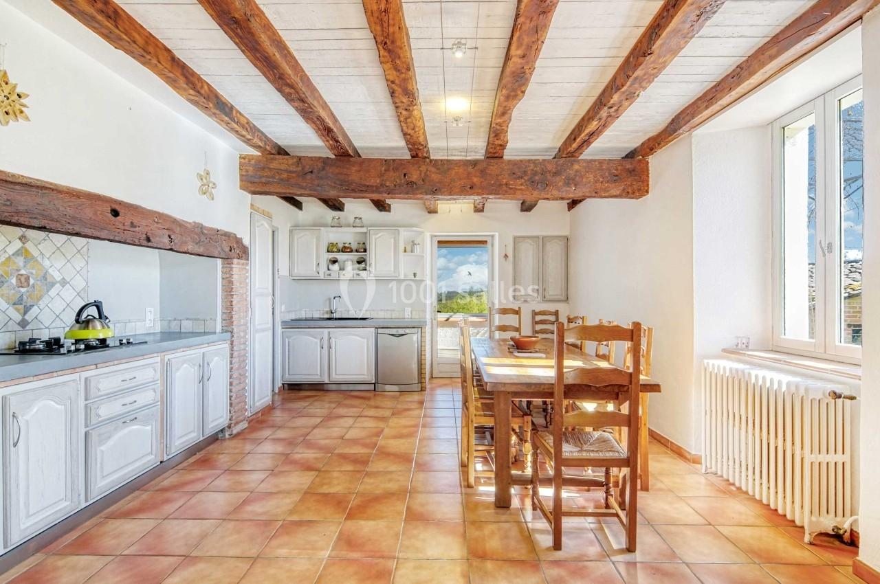 Cuisine lumineuse avec poutres apparentes, carrelage au sol, table en bois et vue sur l'extérieur par une porte vitrée.