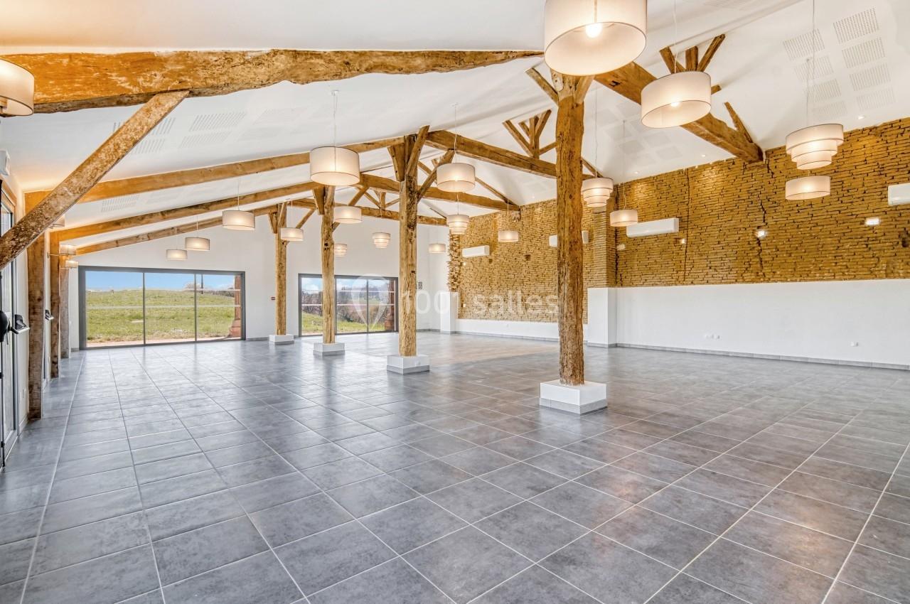 Grande salle lumineuse avec poutres apparentes, murs en briques et carrelage gris, donnant sur un espace extérieur.